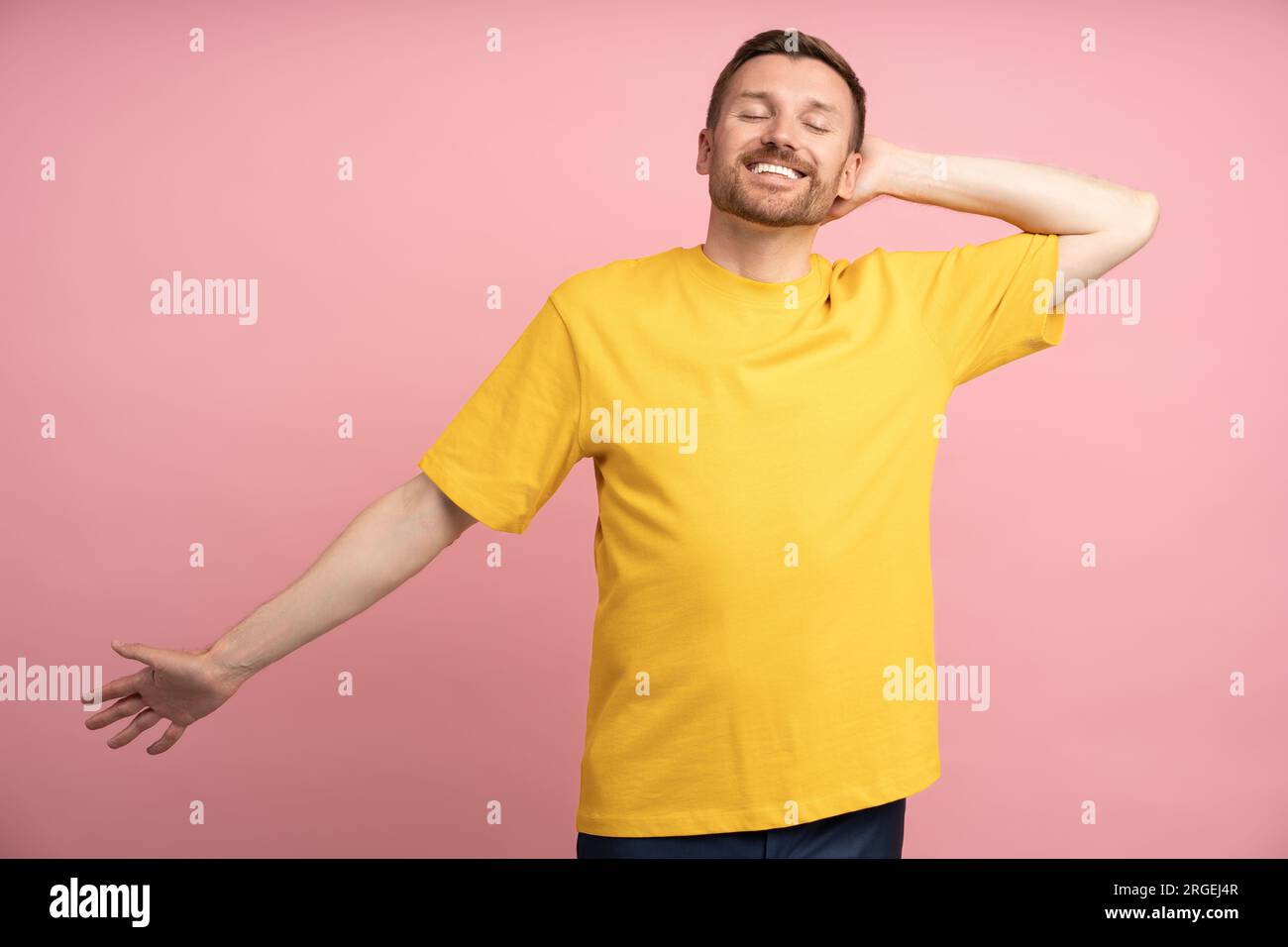 Uomo sorridente spensierato con occhi chiusi e braccio allungato al mattino, parete rosa studio Foto Stock