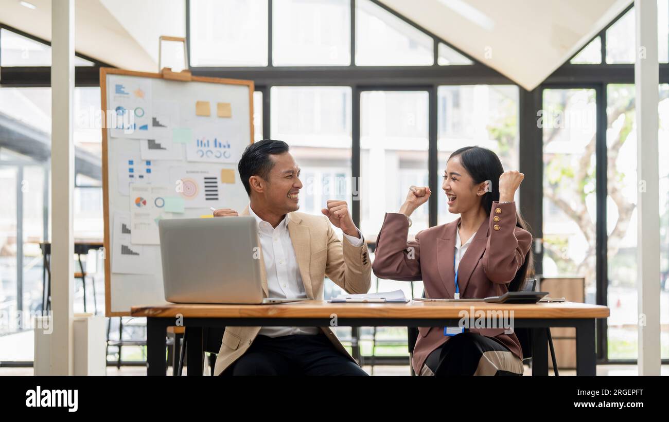 Un allegro uomo d'affari asiatico sta mostrando i suoi pugni e festeggiando un progetto di successo con una collega donna a un tavolo in ufficio. Foto Stock