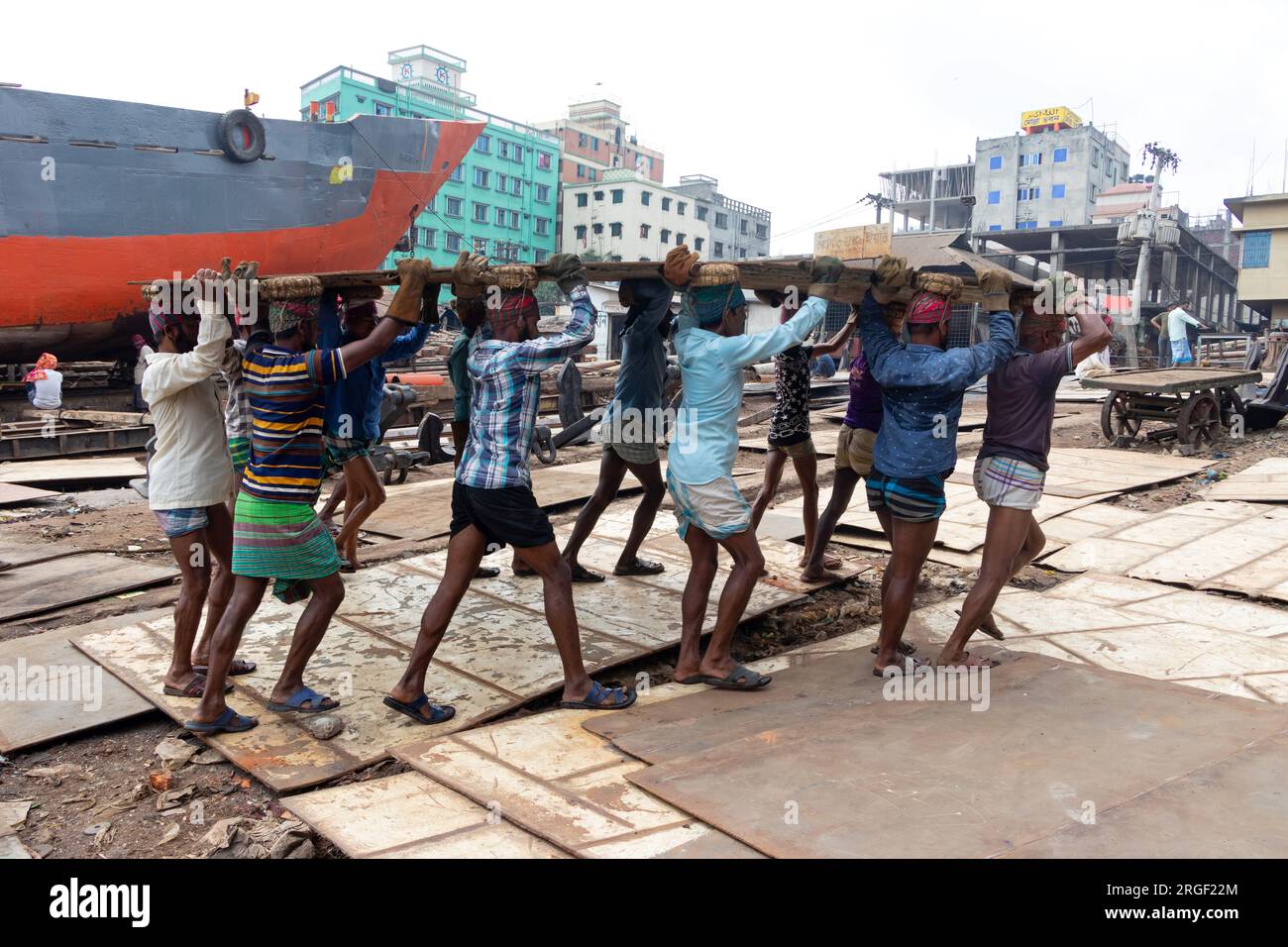 Dhaka, Dhaka, Bangladesh. 9 agosto 2023. Un gruppo di portatori trasporta una pesante piastra di acciaio di 250 kg in un cantiere navale sulla riva del fiume Buriganga, a Keraniganj, vicino a Dacca, Bangladesh. Dozzine di cantieri che occupano 30,96 acri della costa di Buriganga sono stati in funzione negli ultimi 50 anni. Viene utilizzato principalmente per riparare e riparare vecchie navi e costruire nuove navi. Le maestranze lavorano nel cantiere senza caschi, maschere facciali o scarpe di sicurezza. Lavorano sodo tutto il giorno, ma sono ancora pagati salari minimi. Con un numero crescente di ordini da parte di acquirenti sia locali che globali, il cantieristico Foto Stock