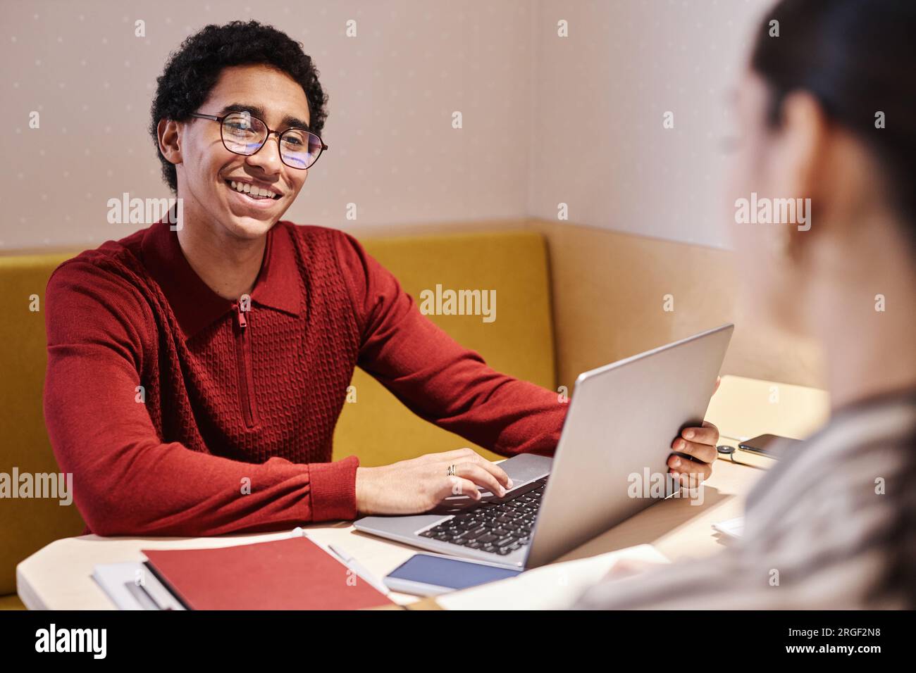 Ritratto di un giovane uomo arabo che indossa occhiali e sorride durante un incontro di lavoro con un collega nello stand dell'ufficio con illuminazione elettrica Foto Stock