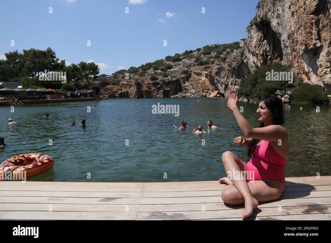 Vouliagmeni Atene Grecia donna che si prende un Selfie sul suo smartphone e turisti nuotare nel lago Vouliagmeni una spa naturale - una volta era una caverna ma il Foto Stock