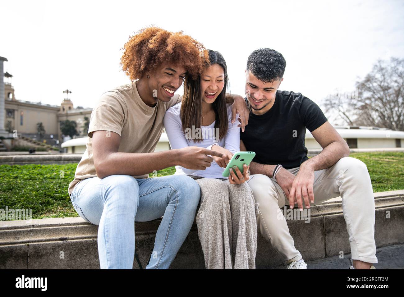 Tre giovani persone diverse che guardano uno smartphone seduto in un parco. Due giovani che sorridono e puntano al telefono di una donna felice. Foto Stock