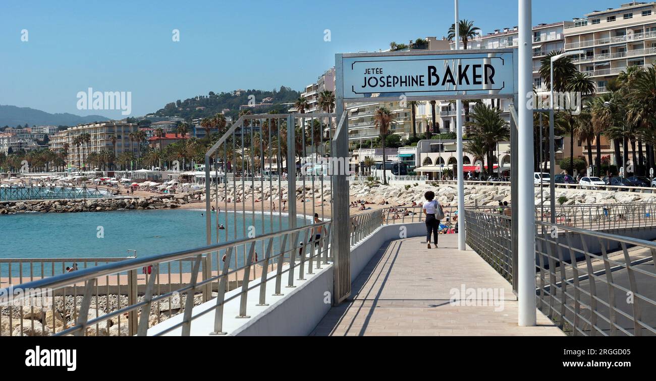 Una donna passeggia lungo Josephine Baker Jetty, Cannes. La spiaggia pubblica del Porto Vecchio di Cannes è a sinistra; gli hotel si affacciano sullo sfondo della Baia di Cannes Foto Stock