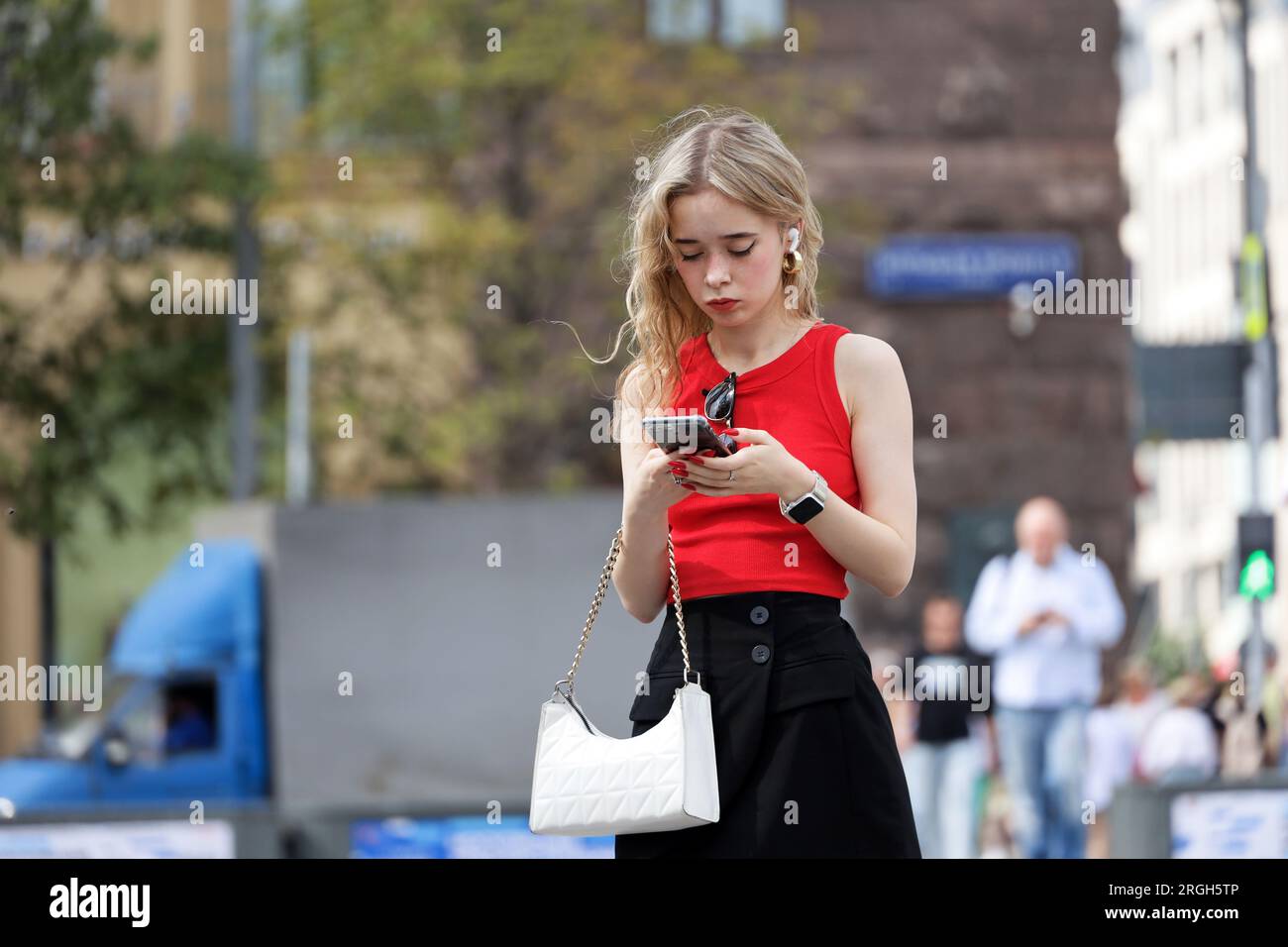 Ragazza attraente con capelli ricci biondi in piedi con lo smartphone in mano in strada, che usa il telefono cellulare nelle città estive Foto Stock