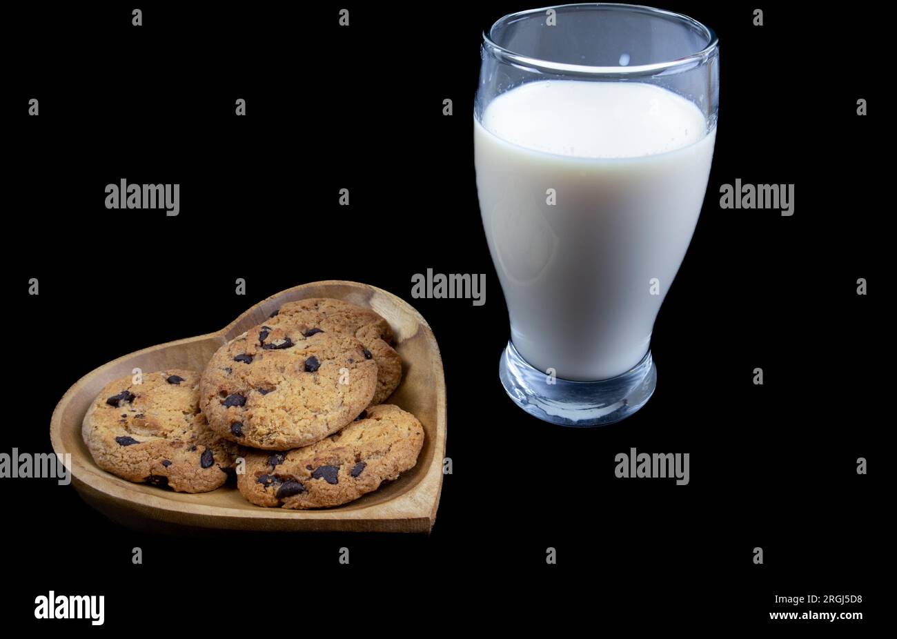 Biscotti con gocce di cioccolato su un piatto a forma di cuore con un bicchiere di latte fresco su sfondo nero Foto Stock