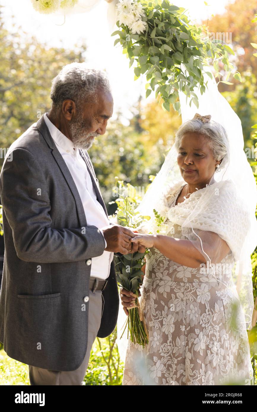 Felice sposo birazziale senior che mette un anello sul dito della sposa alla soleggiata cerimonia nuziale all'aperto Foto Stock