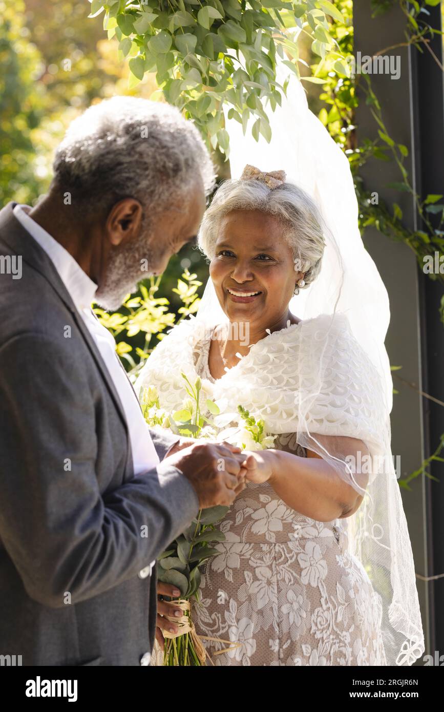 Felice sposo birazziale senior che mette un anello sul dito della sposa alla soleggiata cerimonia nuziale all'aperto Foto Stock