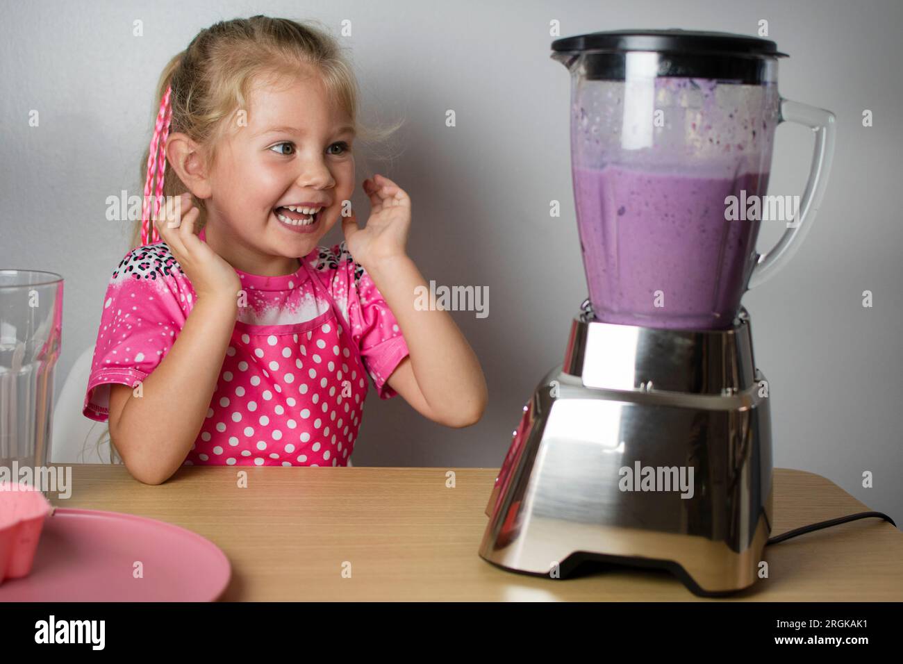 Il bambino Happy Caucasian cute è divertito dal rumore forte del frullatore. Il bambino prepara il frullato. Ragazza che cucina frullato Foto Stock