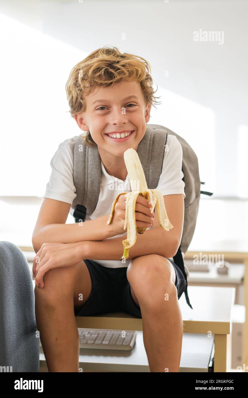 Bambino di scuola allegro con zaino seduto sul tavolo in classe e mangiare banana durante la pausa mentre si guarda la macchina fotografica Foto Stock