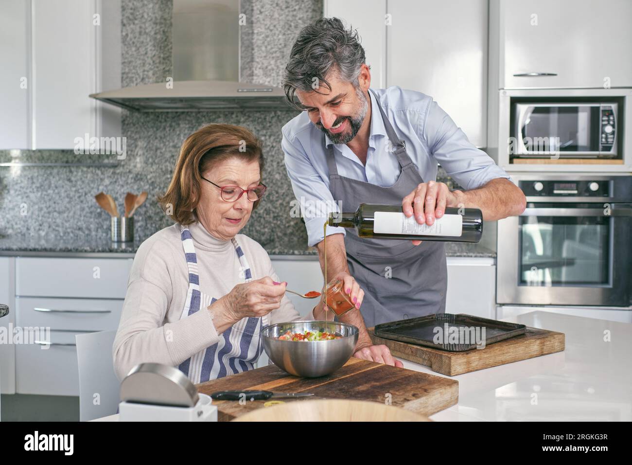 Figlio positivo nel grembiule che versa olio d'oliva nel recipiente con ripieno durante la cottura con la madre vicino al tavolo in cucina leggera Foto Stock