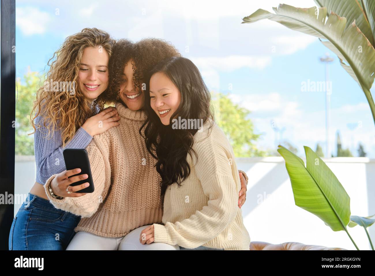 Contenuti giovani donne diverse con maglioni seduti l'uno vicino all'altro e abbracciati mentre si scatta un autoritratto sul telefono cellulare Foto Stock