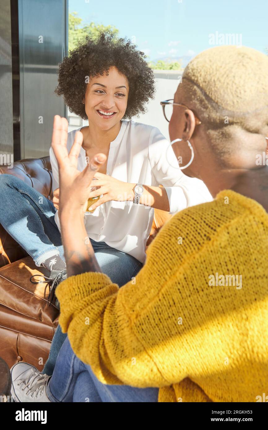 Amiche afro-americane positive che sorridono e si guardano mentre si siedono vicino alla finestra al caffè Foto Stock