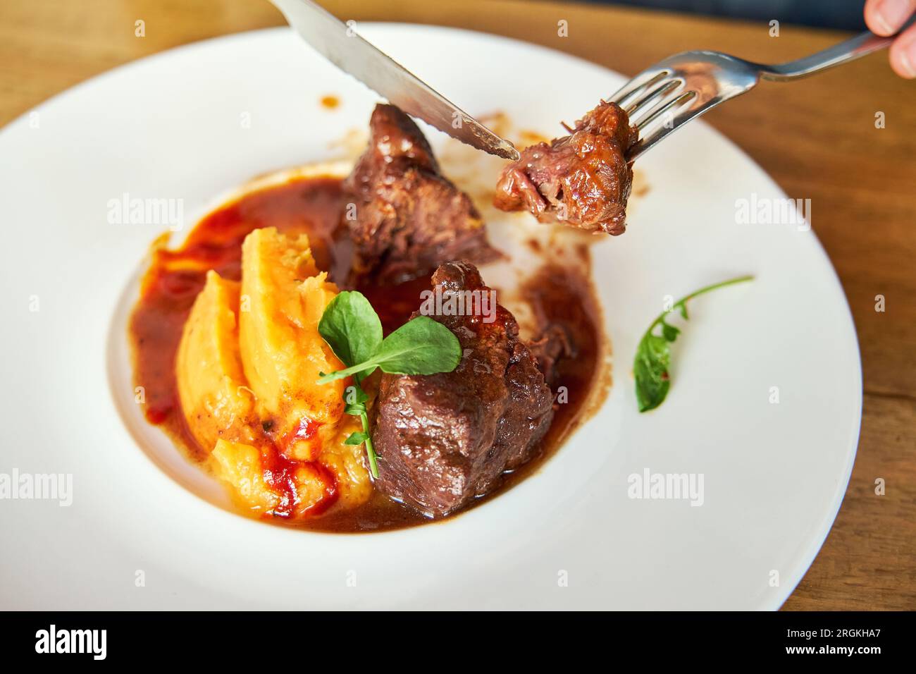Dettaglio delle forchette che consentono a una persona di afferrare un pezzo di carne tagliata da un piatto di guance di vitello con mojo di cocco e purea di tonno con salsa d'arancia Foto Stock