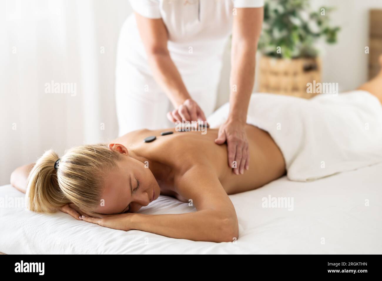 Una donna bellissima si sta godendo un rilassante massaggio alla schiena nel centro termale di cosmetologia. La massaggiatrice applica pietre laviche alla colonna vertebrale. Foto Stock