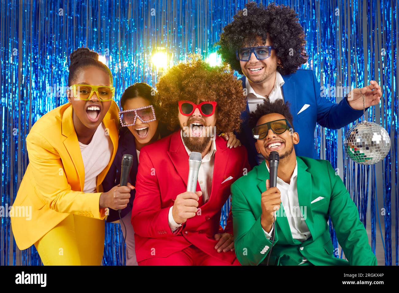 Divertente gruppo di giovani che cantano nei microfoni a una festa, vestiti con costumi luminosi e divertenti bicchieri multicolori, cantando canzoni da pop Foto Stock