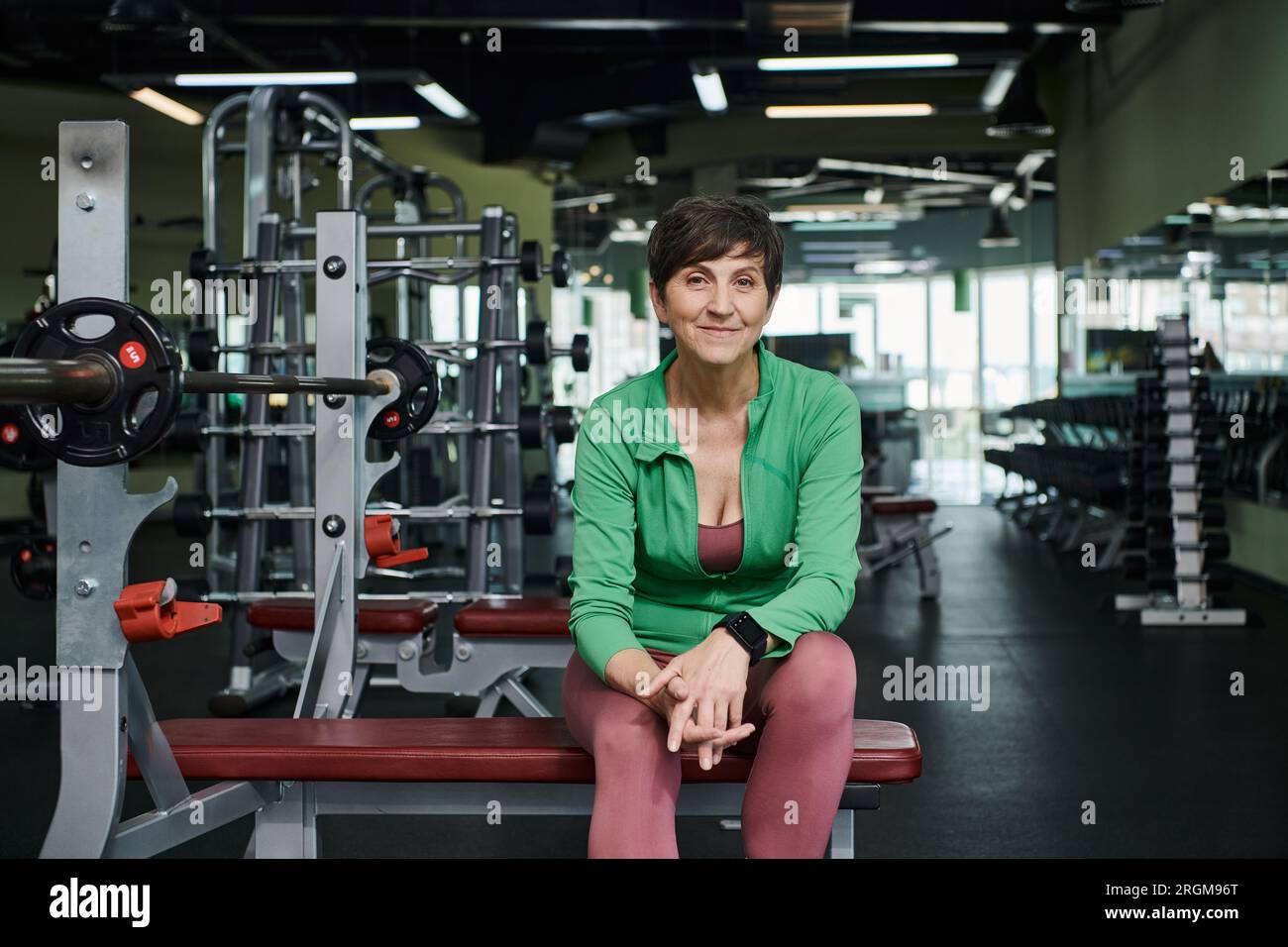 donna anziana felice sorridente e seduta vicino al campanello, ai pesi, all'allenamento in palestra, alla motivazione Foto Stock