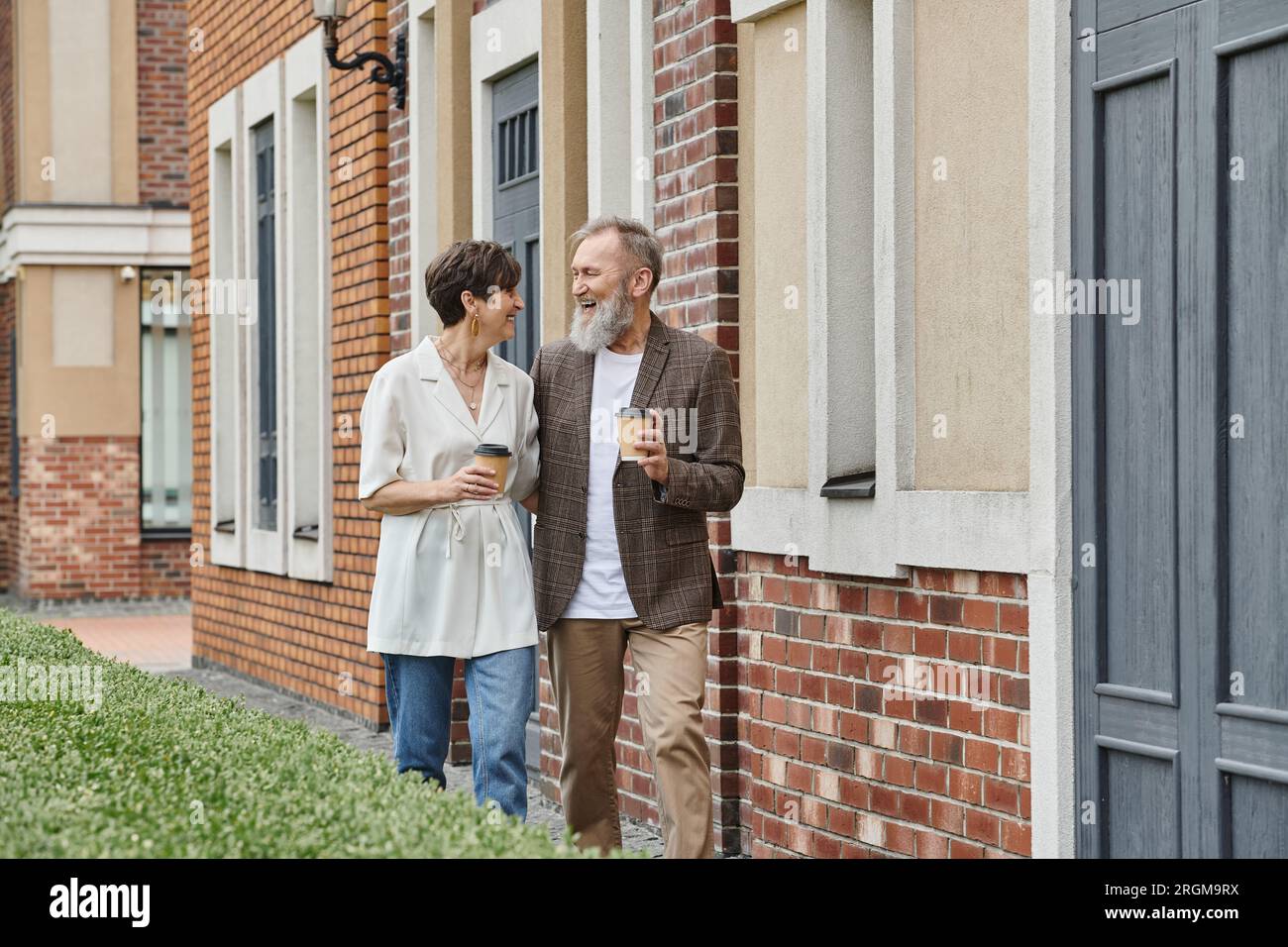 felice coppia anziana che tiene un caffè da andare, camminando vicino all'edificio, uomo e donna anziani, risate Foto Stock