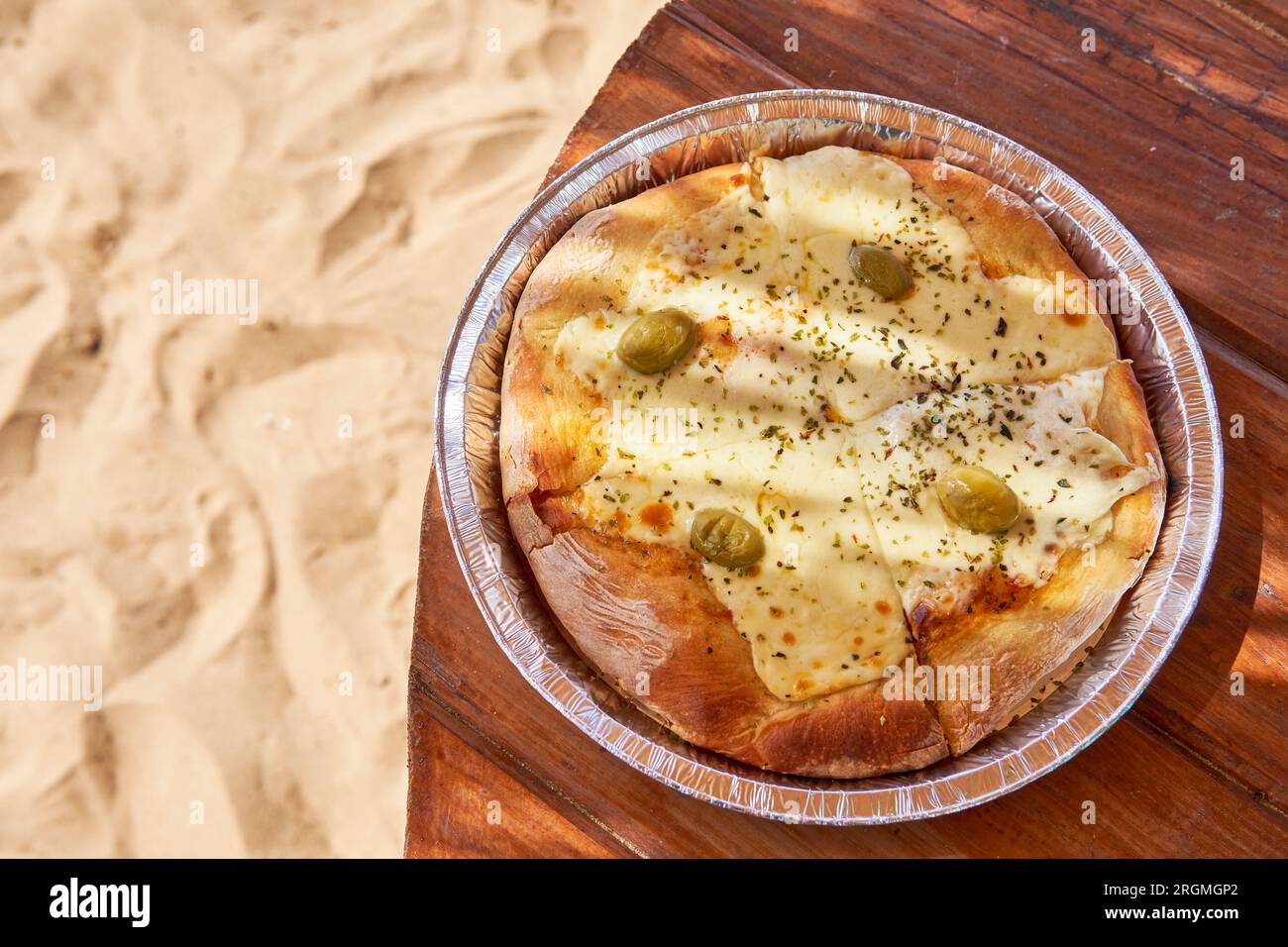 pizza con mozzarella e olive su un vassoio di alluminio su un tavolo di legno sulla sabbia. Fast food per godersi una giornata estiva in spiaggia. Copia spazio Foto Stock