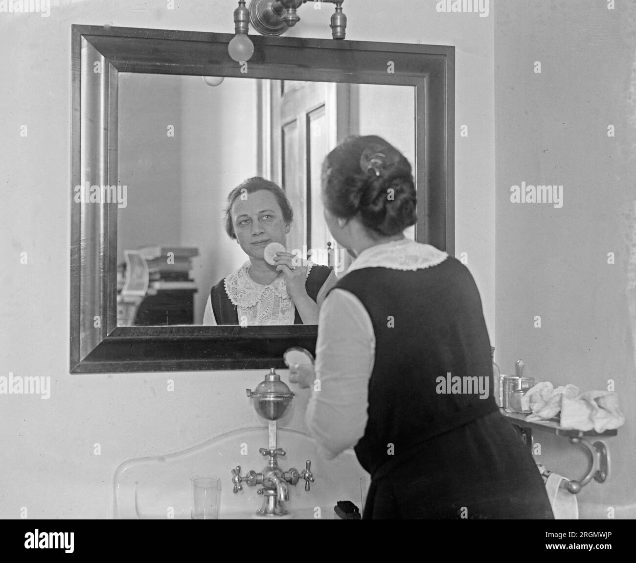 Donna che guarda in uno specchio del bagno che applica trucco a polvere CA. 1923 Foto Stock