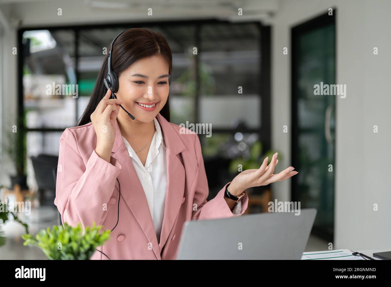 Una bella operatore asiatica di call center con una cuffia sta conversando con un cliente su un computer portatile, lavorando in un ufficio di call center. Foto Stock