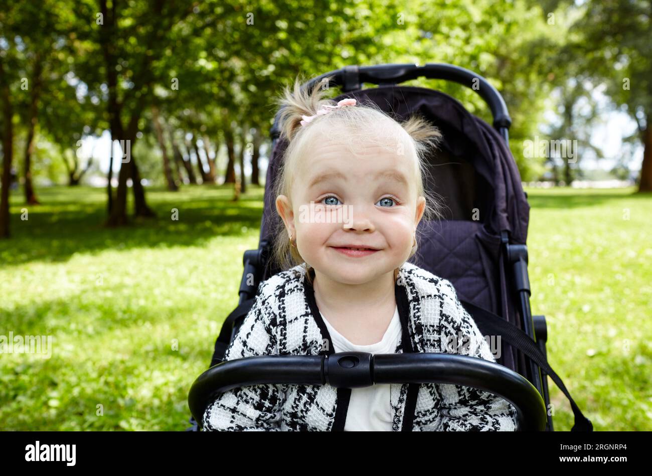 Bambino in passeggino su una passeggiata nel parco estivo. Adorabile bambina seduta su un passeggino, divertente espressione facciale. Bambino in buggy Foto Stock