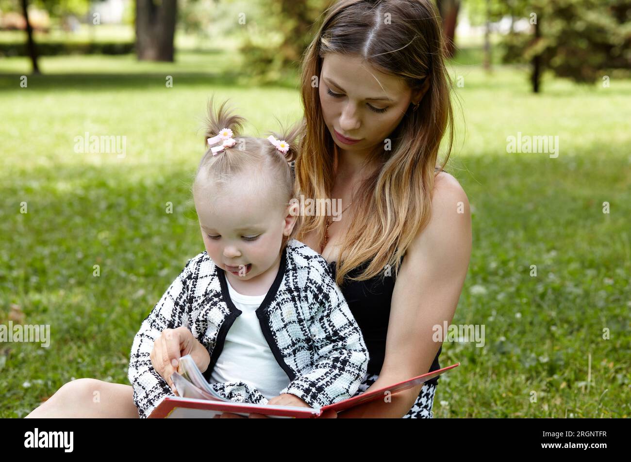 Madre e figlia sedute sull'erba nel parco cittadino estivo e leggono un libro. Concetto di infanzia, tempo libero e persone: Buon riposo in famiglia e buon divertimento Foto Stock