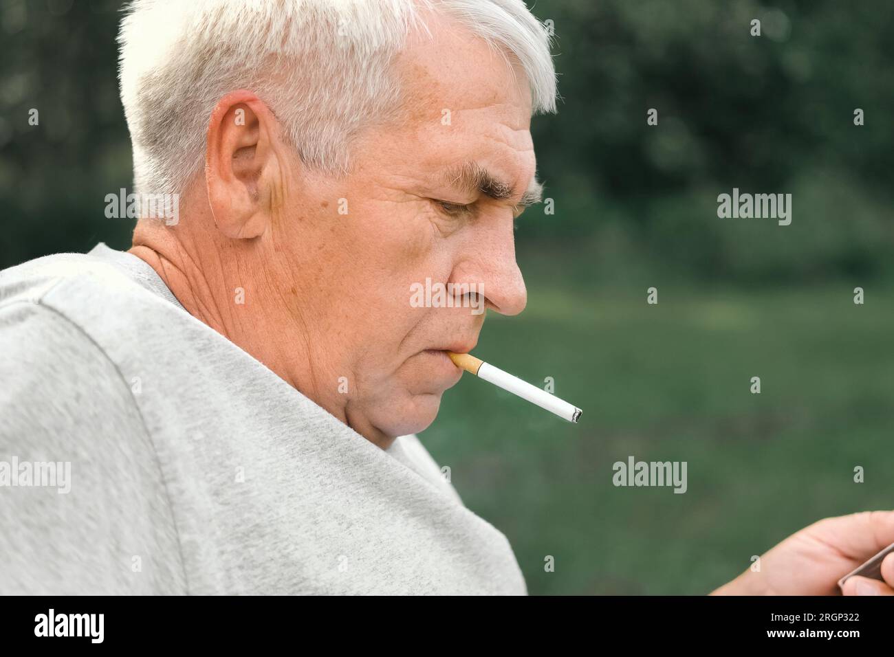 Uomo anziano che accende una sigaretta all'aperto su sfondo verde. Vecchio uomo premuroso che fuma, guardando da parte. Faccia da vicino. Pensione rilassata Foto Stock