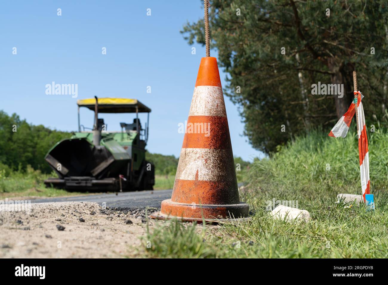 Cono stradale nel cantiere di una nuova strada. Attrezzatura finitrice in background. Macchina per pavimentazione cingolata per la posa di asfalto. Foto Stock