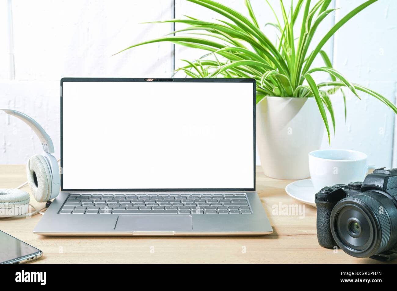 Apri il computer portatile con uno schermo bianco vuoto, fotocamera, cuffie e una pianta sulla scrivania di un giornalista o fotografo contro una parete dipinta di bianco, finta u Foto Stock