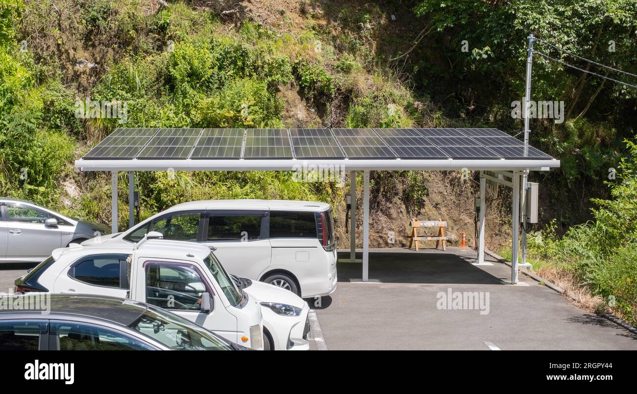 Una stazione di ricarica coperta per veicoli elettrici con pannelli solari sul tetto in un parcheggio dell'hotel pieno di veicoli con motore a combustione in una calda estate Foto Stock