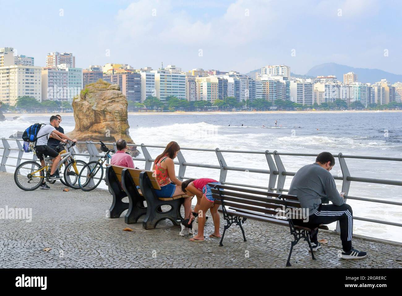 Niteroi, Brasile, gente seduta in una panchina del quartiere sul lungomare. Le onde del mare si schiantano contro una grande roccia. Il paesaggio urbano o lo skyline si trovano nel ba Foto Stock