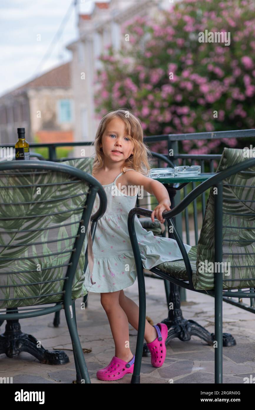 Una bambina carina con un vestito e sandali rosa a un tavolo in una caffetteria in un giorno d'estate. Un caffè, una terrazza estiva, una bambina vicino a un tavolo con sedie. Foto Stock