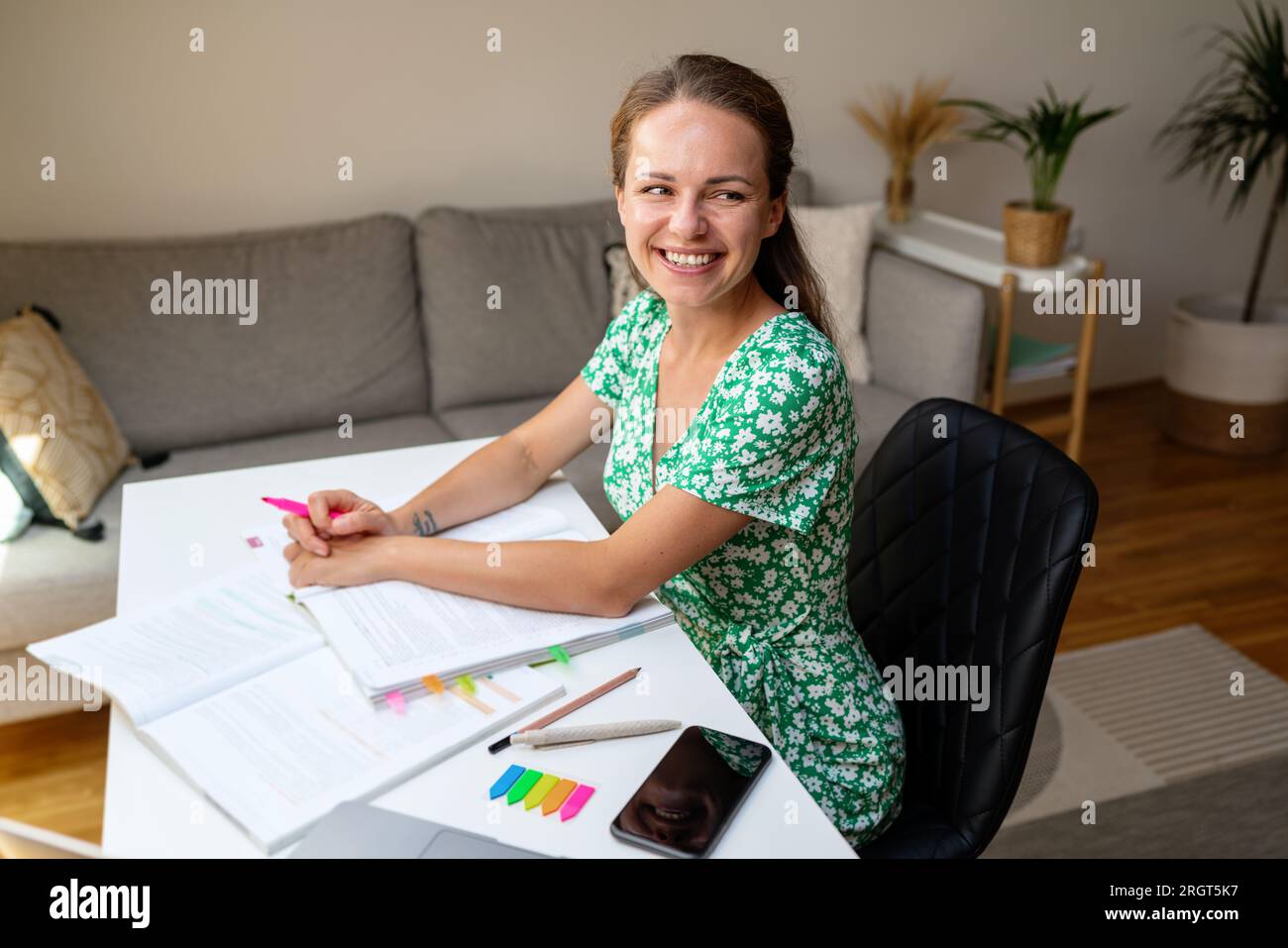 una giovane studentessa bruna che indossa un vestito estivo si siede alla scrivania a casa facendo i compiti e sorridendo. Foto Stock