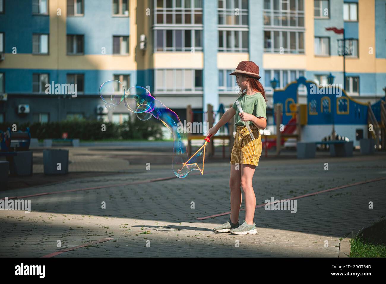 Una graziosa ragazza in età scolare soffia grandi bolle di sapone nel cortile di un edificio a più piani in estate. concetto estivo, giochi all'aperto Foto Stock