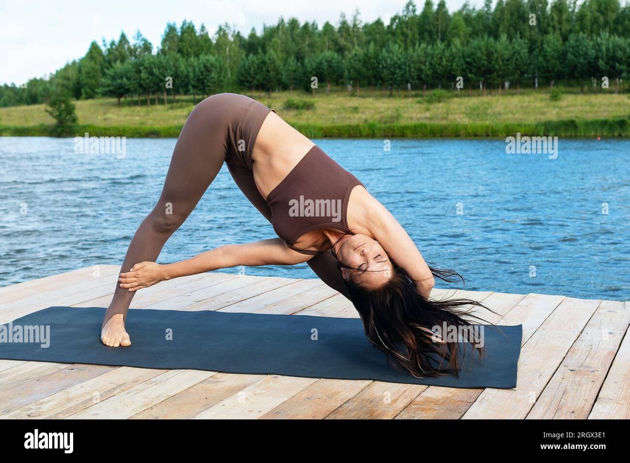 Una donna che pratica yoga, esegue una variante dell'esercizio Trikonasana, posa triangolare, si allena in abbigliamento sportivo sulla riva di un lago in una calda estate Foto Stock