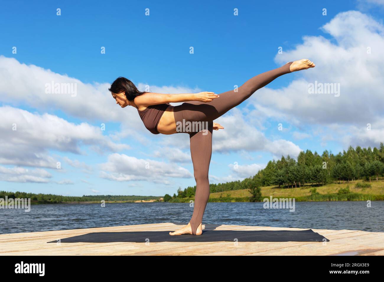 Una donna che pratica yoga, facendo una variazione dell'esercizio Virabhadrasana, la posa numero tre guerriera, si sta allenando in abbigliamento sportivo sulla riva di un Foto Stock