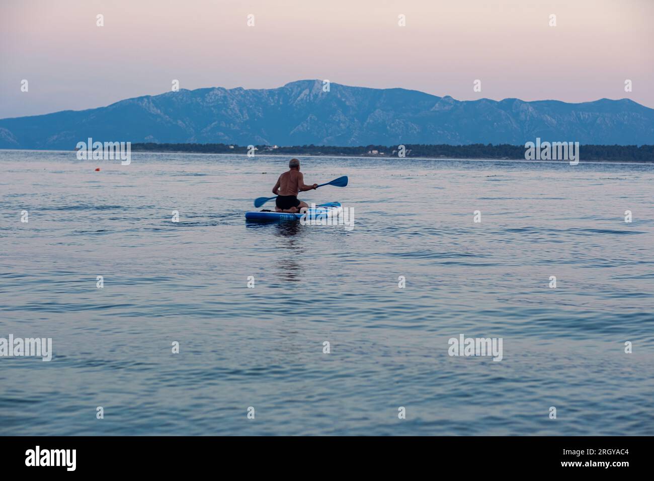 L'uomo dai capelli grigi e dalle barbabietole invecchiate nuota su una linfa in mare al tramonto. Bella e tranquilla pulizia del mare e delle montagne sullo sfondo. Foto Stock