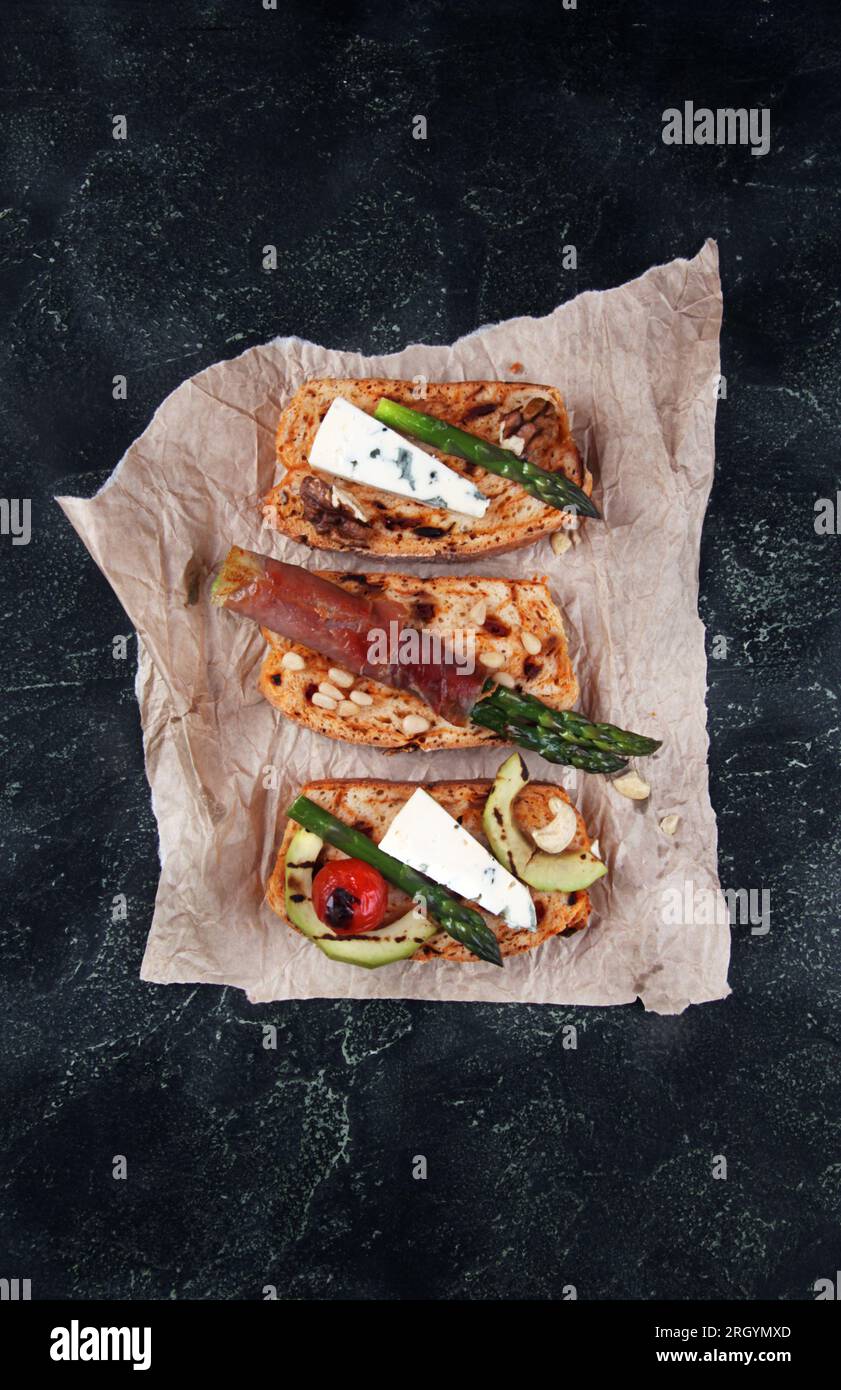 Pane tostato fatto in casa servito con asparagi verdi avvolti in pancetta, formaggio, fette di avocado, ciliegia di pomodoro alla griglia e noci su fondo di cemento scuro Foto Stock