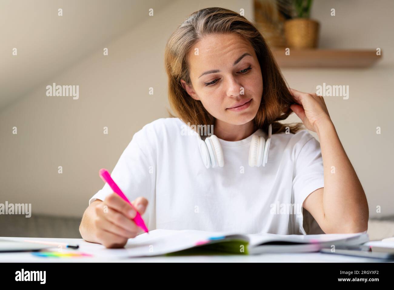 donna bruna con le cuffie sul collo, indossa una maglietta bianca seduta al tavolo a casa e fa i compiti usando il libro degli esercizi dello studente e il rosa f Foto Stock