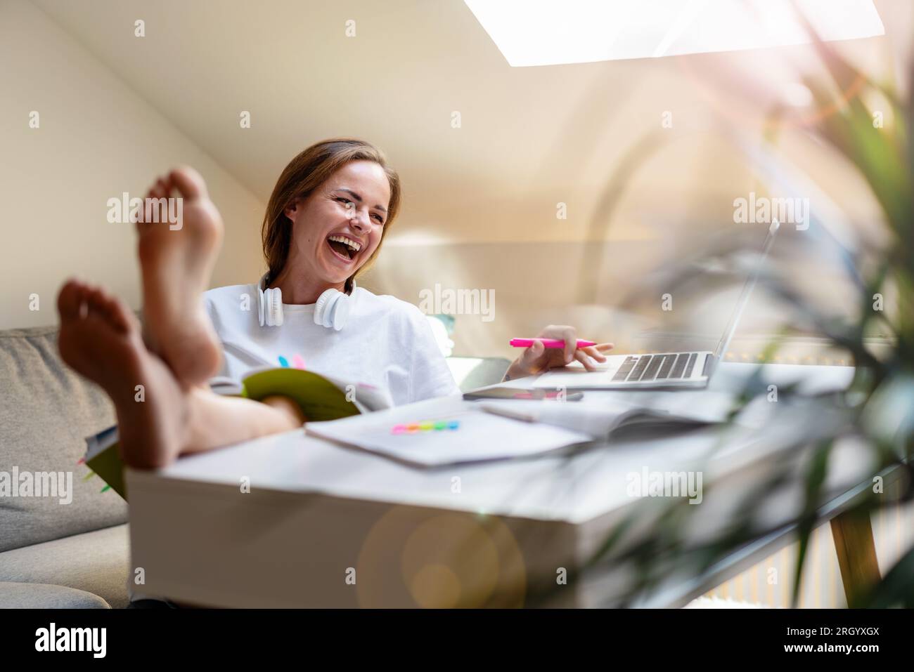 Una giovane studentessa siede alla scrivania di casa con i piedi in alto sul tavolo e si diverte, ridendo mentre usa il suo notebook. Foto Stock