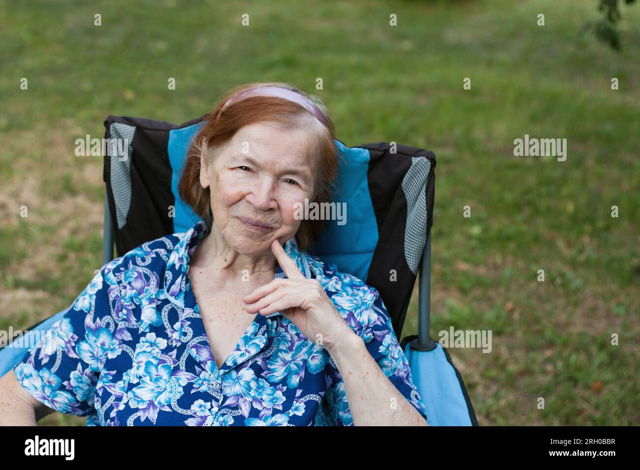 Ritratto della nonna anziana sorridente in giardino a casa. l'anziana si sta riposando seduto sulla sedia, guardando la macchina fotografica, il frutteto sullo sfondo. Somma Foto Stock