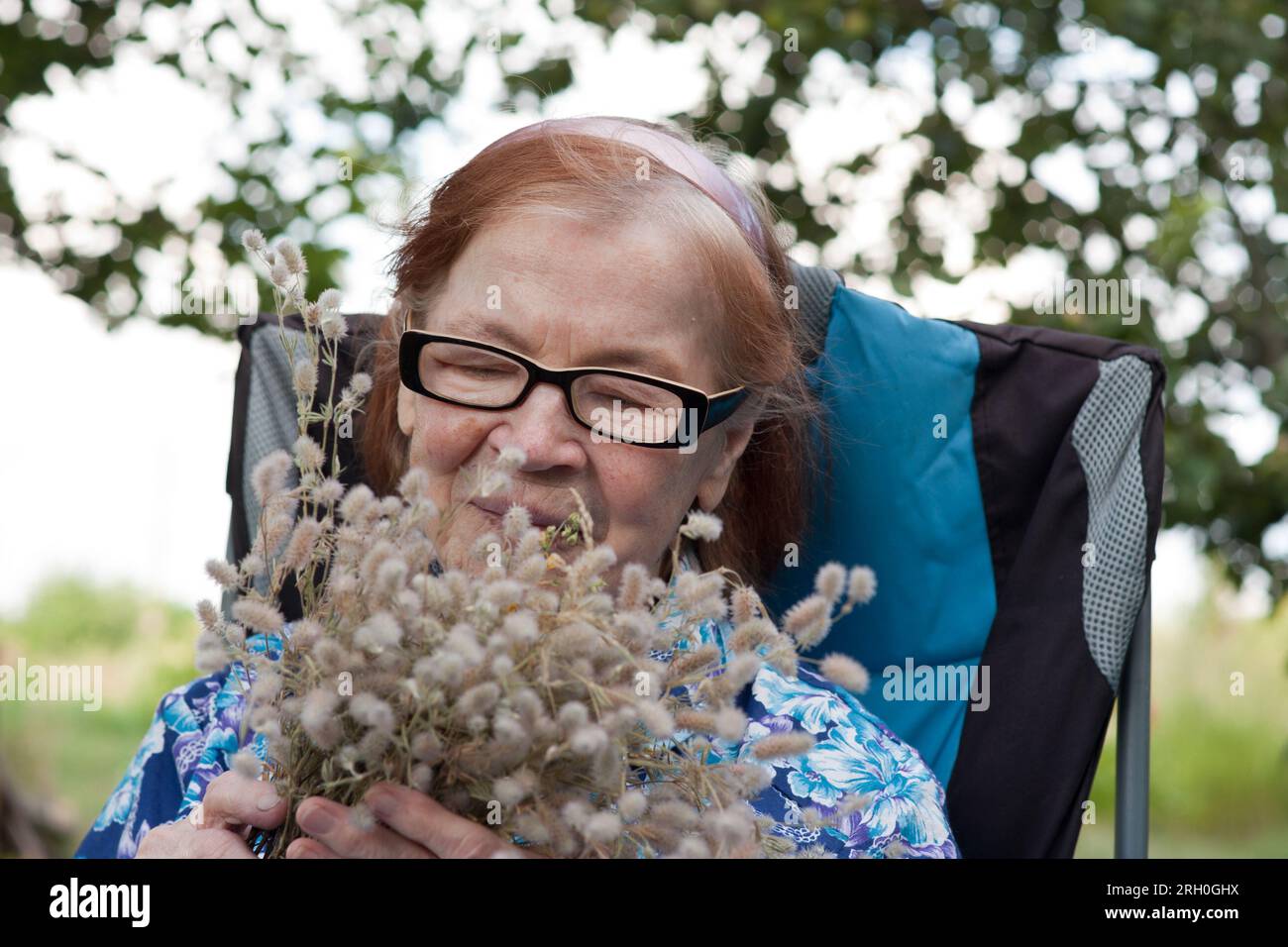 Ritratto della nonna anziana felice in giardino. anziana sopra i 80 anni con bouquet di fiori selvatici, riposa, seduta in sedia, nella natura. Nonni Day. Foto Stock