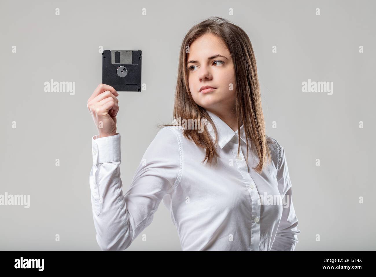 donna con capelli di media lunghezza, vestita in modo ordinato di bianco, tiene un floppy disk nero, osservandolo mentre contempla il rapido progresso della tecnologia Foto Stock