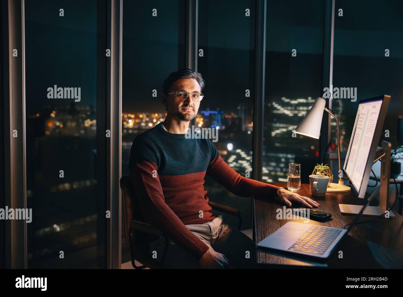 Un imprenditore maschio caucasico lavora fino a tardi alla sua scrivania, conducendo ricerche di mercato con il suo portatile. Si siede con sicurezza, guardando la telecamera, S Foto Stock