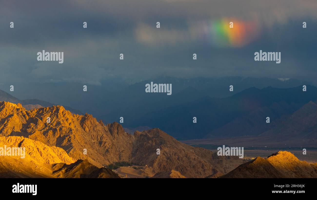 Raggi del sole che cadono su montagne aride con nuvole e un arcobaleno nel cielo Foto Stock
