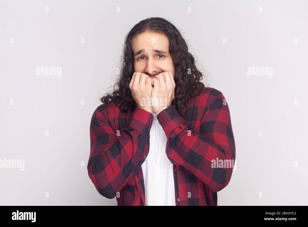 Ritratto di uomo barbuto in camicia rossa a scacchi sembra nervoso, morde unghie da dito, guarda film in movimento, anticipa quale sarà la fine. Riprese in studio in interni isolate su sfondo grigio. Foto Stock