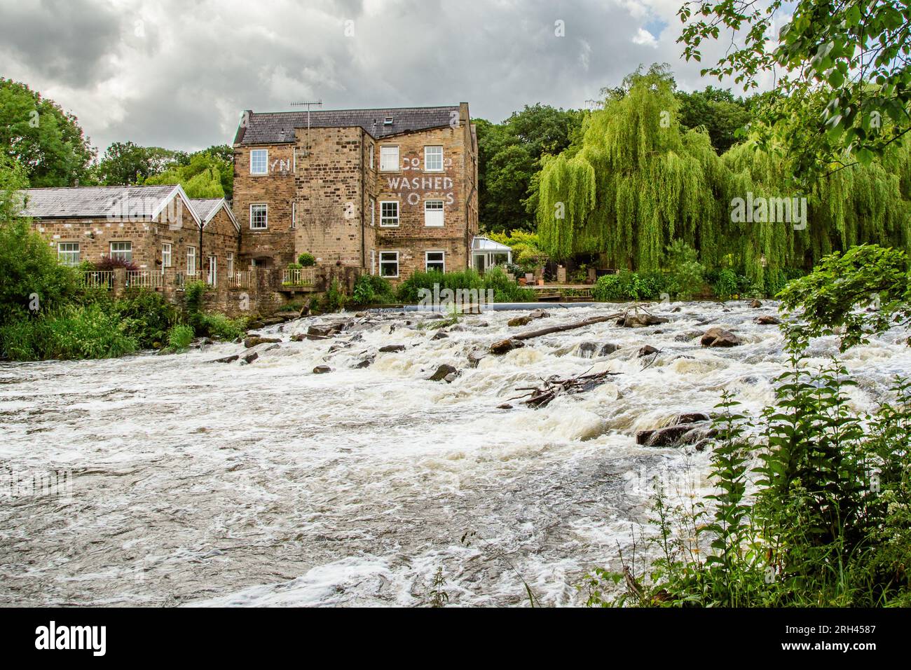 Il fiume Aire che scorre su una diga a Baildon, nello Yorkshire. L'Hirst Mill (un ex mulino per il mais) si vede dall'altra parte dell'acqua. Foto Stock