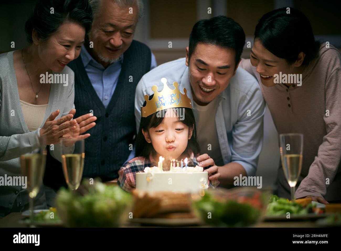 piccola ragazza asiatica che soffia candele mentre la famiglia di tre generazioni festeggia il suo compleanno a casa Foto Stock
