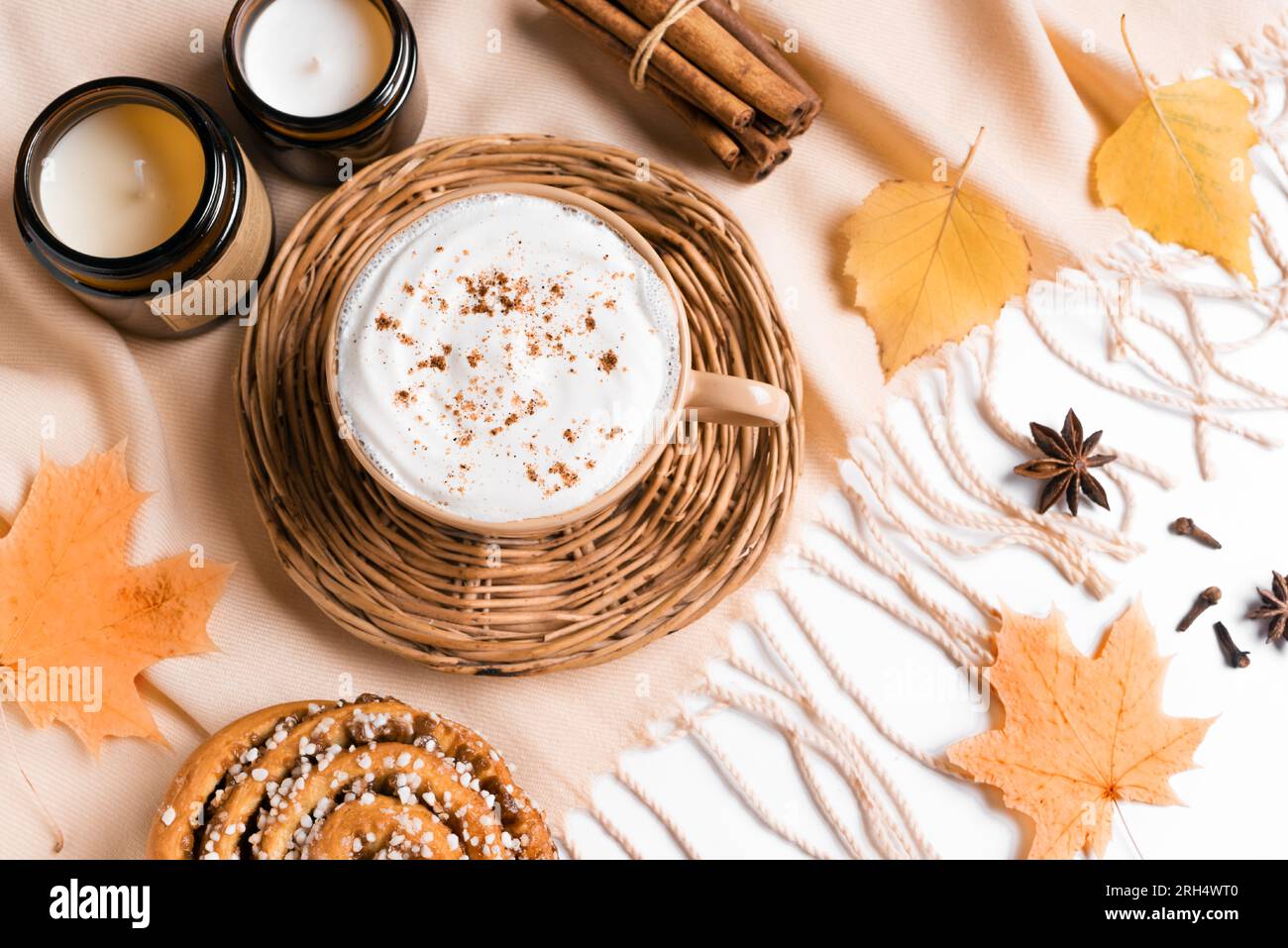 Concetto estetico autunnale. Tazza di caffè cappuccino con cinnabon, candele, quadri beige e foglie autunnali su sfondo bianco da vicino. Foto Stock