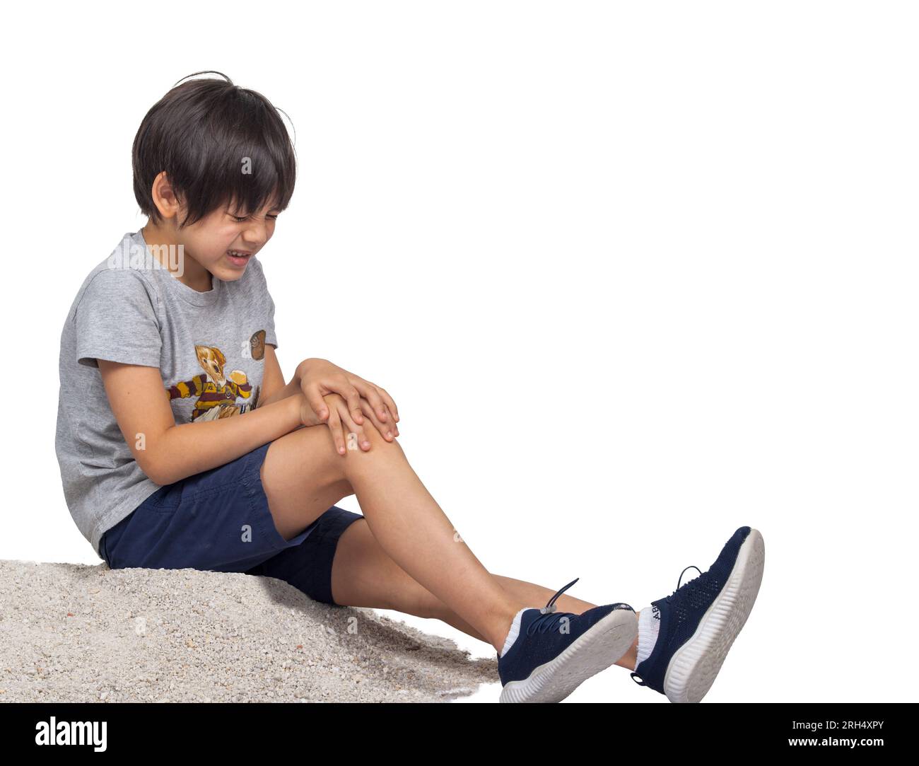 Foto in studio di un ragazzo panasiatico seduto sulla sabbia, che piange su un ginocchio ferito isolato su uno sfondo bianco, mostrando vulnerabilità ed emozione. Foto Stock
